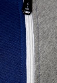 Tessuto blu e grigio con zip, caratterizzato da una cerniera bianca e accessori neri. Dettaglio della cucitura testurizzata visibile lungo le cuciture.