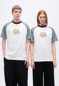 Άσπρα μπλουζάκια με κοντά μανίκια σε μπλε και σκούρο μωβ μοτίβο, με κίτρινο κεντημένο λογότυπο στο μπροστινό μέρος και ριμπ λαιμόκοψη.