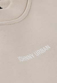 Sudadera beige con un cuello de canalé y texto en blanco "JOHNNY URBAN" impreso en la parte frontal. Textura de tela suave, diseño casual.
