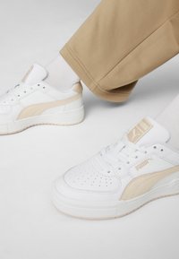 Valge sneaker, millel on sileda nahaga pealis, beež aktsent, ümar varvas, perforatsiooni muster ja tekstureeritud kummitald.