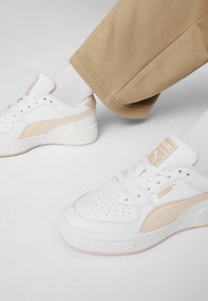 Zapatilla blanca con un upper de cuero suave y un acento beige, punta redonda, diseño perforado y suela de goma texturizada.