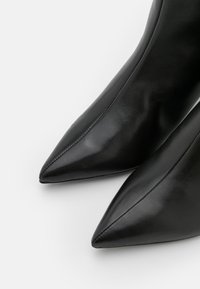 Botas de tobillo de cuero negro con punta afilada y textura suave, que presentan costuras limpias y un diseño elegante y aerodinámico.
