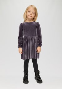 Paarse fluwelen jurk met lange pofmouwen en een geplooide rok, gecombineerd met zwarte leggings en glanzende zwarte veterschoenen.