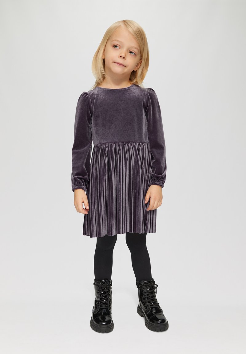 Paarse fluwelen jurk met lange pofmouwen en een geplooide rok, gecombineerd met zwarte leggings en glanzende zwarte veterschoenen.