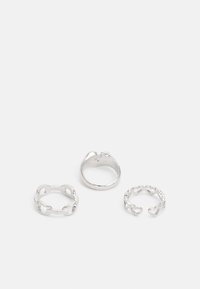 Trois anneaux en argent : un bandeau lisse, un avec un design de chaîne entrelacée, et un avec des découpes en forme de cœur. Fini en métal poli.