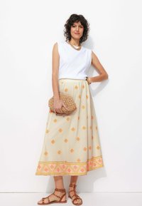 Jupe beige avec des motifs géométriques orange et rose, associée à un haut sans manches blanc et des sandales à brides marron ; tenant une pochette texturée.