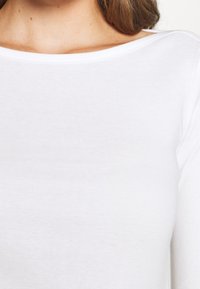 Vit långärmad tröja i mjukt material, med bred halsringning och slät textur, med minimala sömnadsdetaljer längs kanterna.