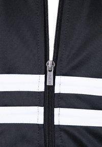 Giaccone nero con zip e strisce orizzontali bianche. Presenta un tirazip metallico con incisione "UNFR". Tessuto testurizzato e durevole.