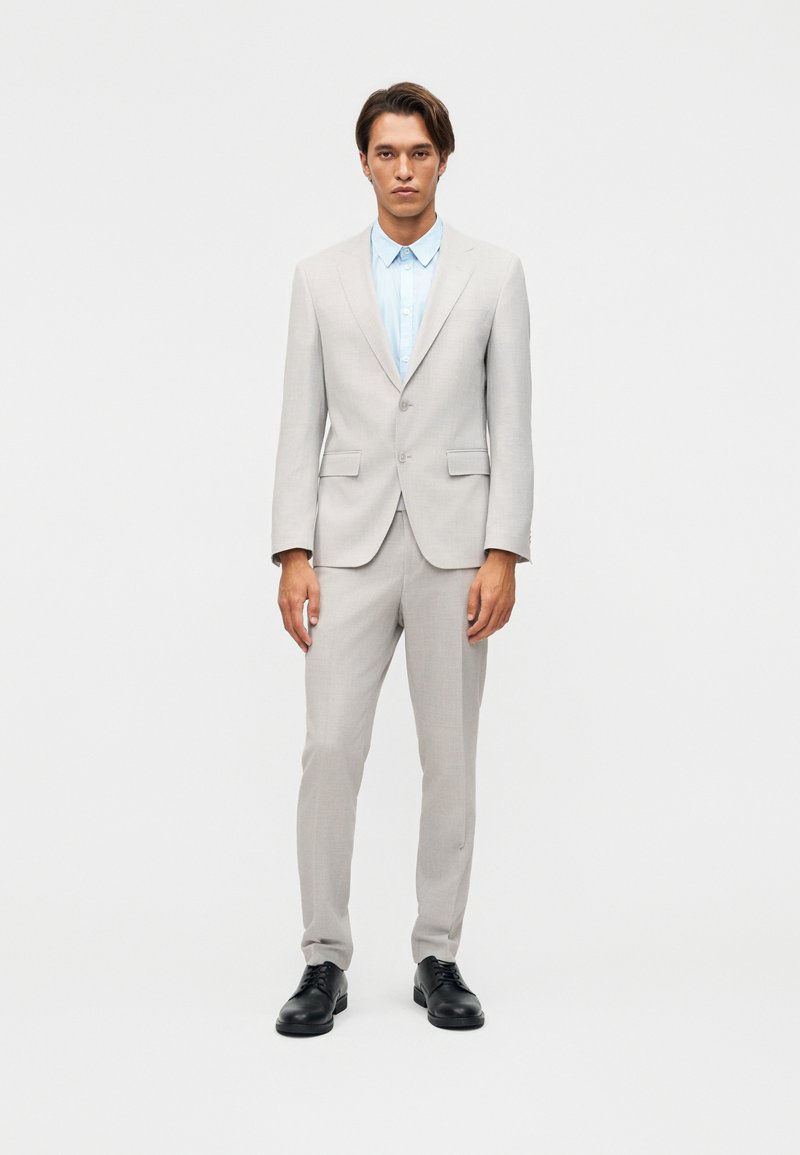 Homme debout, face à l'avant, portant un costume gris clair, une chemise bleu clair et des chaussures de ville noires, sur un fond blanc uni.