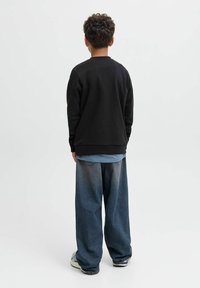 Sudadera negra con cuello redondo, combinada con una camisa de color azul claro y pantalones vaqueros de gran tamaño. Usada con zapatillas deportivas gris y blancas.
