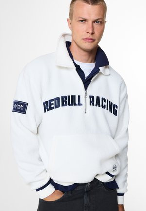 Joven con una chaqueta de forro polar blanca de Red Bull Racing con cremallera, de pie con una mano en el bolsillo frente a un fondo liso.