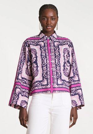 Femme portant une chemise boutonnée à motifs violets et roses avec des manches larges et un pantalon blanc, se tenant devant un fond blanc uni.