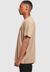 Young man in beige T-shirt with small dripping pizza slice graphic on chest, standing sideways against plain light background.