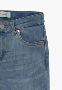 Vaaleansiniset farkut, joissa on kapea malli, näyttävät etutaskustaan, kuparisista niiteistä ja napin kiinnityksestä, sekä näkyvistä ompeluista.