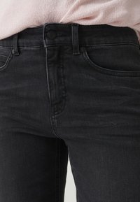 Jeans in denim nero con chiusura con bottoni e cerniera indossati con un top in maglia rosa chiaro infilato.