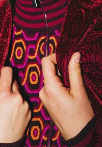 Veste en velours rouge avec texture florale, fermée par une fermeture éclair au col, portée sur un pull à motif géométrique aux tons violet, orange et jaune. Mains agrippant le tissu.