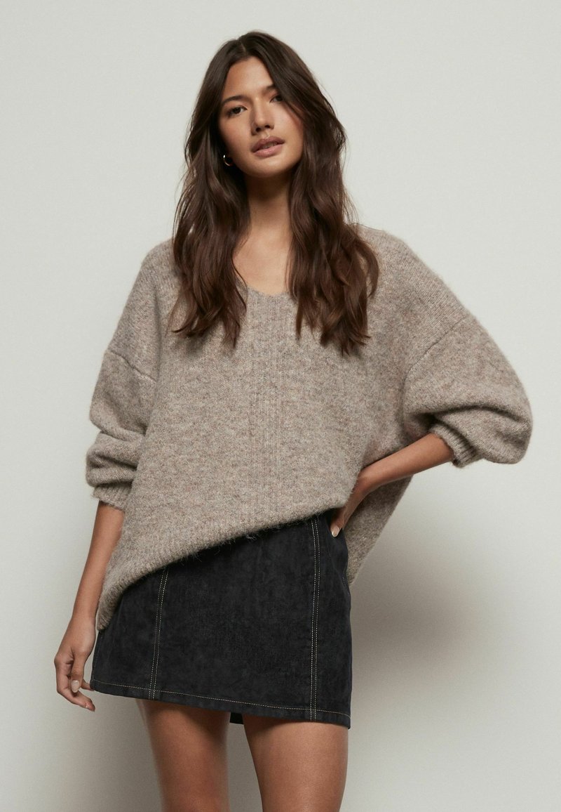 Un pull en tricot beige épais avec un décolleté en V et des manches surdimensionnées, associé à une mini-jupe en daim noire avec des coutures jaunes.
