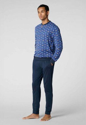 Giovane uomo in piedi a piedi nudi, indossa una camicia blu a maniche lunghe con piccole stampe colorate e pantaloni aderenti blu scuro.