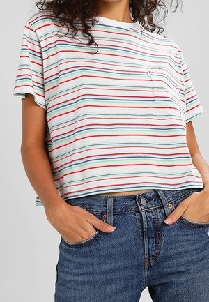 Vrouw draagt een wit crop top met korte mouwen met horizontale rode, groene en blauwe strepen en blauwe jeans met hoge taille, handen in de zakken.
