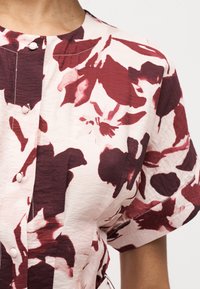 Κοντινή λήψη μπλούζας με κοντά μανίκια και στρογγυλή λαιμόκοψη, κρεμ βάση και μεγάλο σκούρο κόκκινο λουλουδάτο σχέδιο, με κουμπιά καλυμμένα με ύφασμα μπροστά.