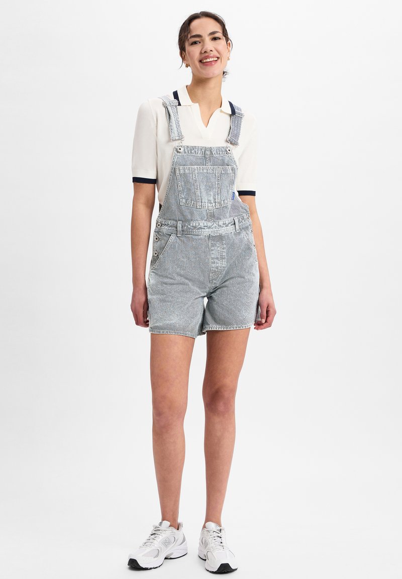 Vrouw die glimlacht, gekleed in een witte polo met korte mouwen en lichtblauwe gestreepte denim short-overall met witte sneakers, staand voor een witte achtergrond.
