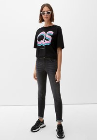 Camiseta negra con un gráfico colorido de "QS", combinada con jeans oscuros de corte slim y zapatillas deportivas negras con acentos blancos.