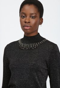 Suéter negro con un cuello alto, que presenta un collar brillante de joyas hecho de cuentas claras y oscuras, sobre una tela texturizada.