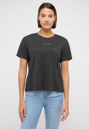 Woman with short brown hair wearing a black Mustang Jeans t-shirt and light blue jeans, standing against a plain light gray background.