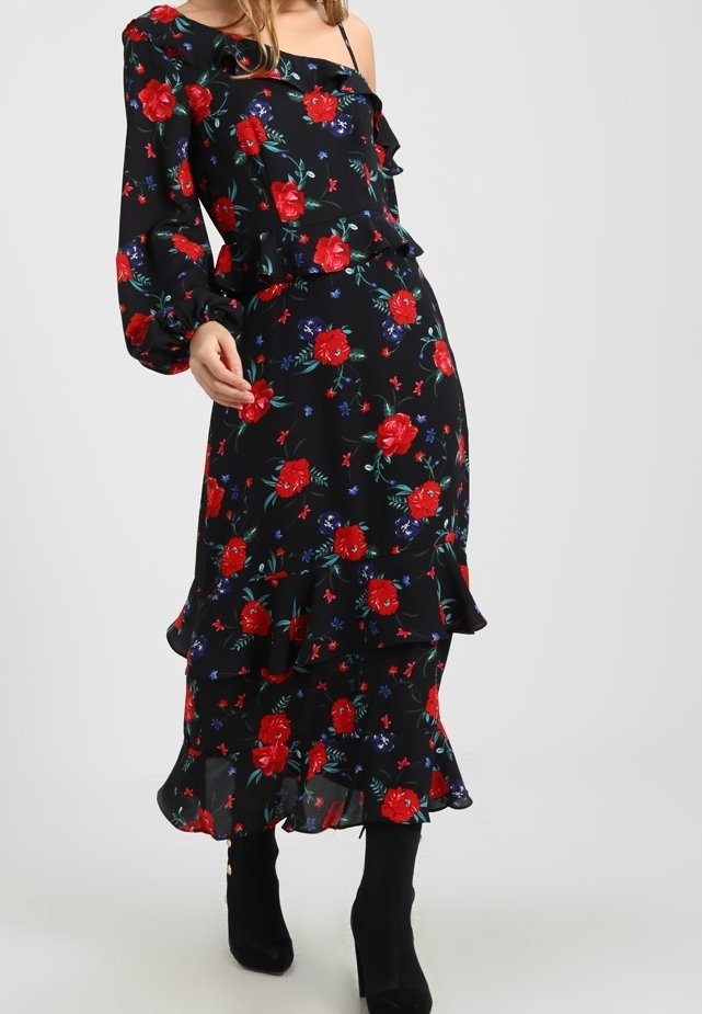 Kvinna i en svart blomstrig klänning med röda och lila blommor, med en lång ärm och volangkant, kombinerad med svarta klackboots.