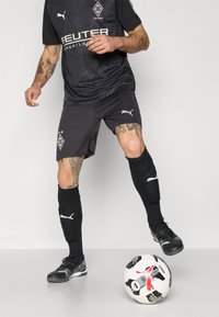 Camiseta deportiva negra de Puma con un patrón de puntos, pantalones cortos grises, calcetines negros y zapatos negros. El jugador está posicionado cerca de un balón de fútbol con patrón.