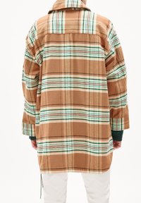 Person som bär en beige rutig långärmad skjorta med gröna och vita ränder, sedd bakifrån, tillsammans med vita byxor.