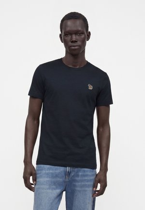 Mand iført en ensfarvet sort T-shirt med en lille farverig zebra-logo på venstre bryst og lyseblå jeans, der står mod en hvid baggrund.