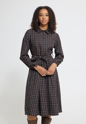 Femme aux cheveux bouclés portant une robe à manches longues, boutonnée, écossaise marron et bleue avec une ceinture à la taille et des bottes marron jusqu'aux genoux.