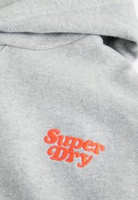 Sudadera gris hecha de una tela suave con cordón. Presenta un logo rojo bordado de 'Super Dry' en la parte delantera. Textura suave, diseño casual.