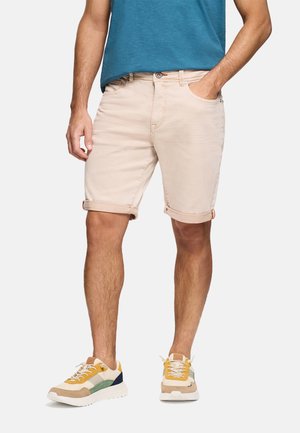 Hombre con pantalones cortos enrollados beige claro, camiseta azul y zapatillas multicolor, de pie con una mano en el bolsillo.