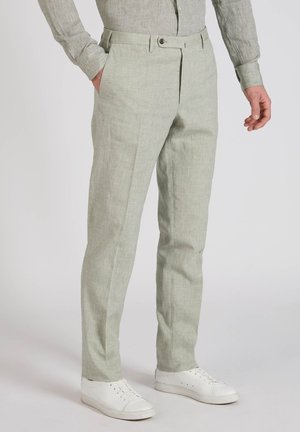 Mężczyzna w jasnoszarych lnianych spodniach, białych trampkach i pasującej, jasnoszarej lnianej koszuli z długim rękawem, stoi z jedną ręką w kieszeni.