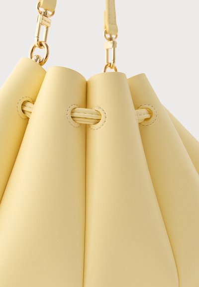 Κίτρινη δερμάτινη τσάντα τύπου κουβά με χρυσά κούμπωμα και λεπτομέρειες κορδονιού, κοντινή φωτογραφία σε υφή υλικού και ραφές.