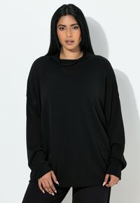 Frau mit langen schwarzen Haaren, die ein lockeres schwarzes Langarmshirt und eine schwarze Hose trägt, steht vor weißem Hintergrund, die Hände entspannt an den Seiten.