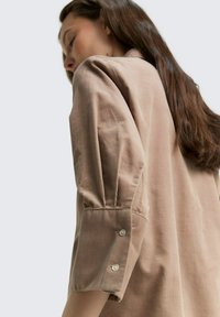 Lichtbruine blouse van textuurstof. Heeft opbollende mouwen en kleine knoopaccenten bij de manchet. Eenvoudig, ontspannen ontwerp.