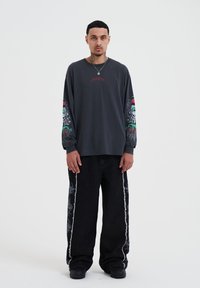 Dunkelgraues Langarmshirt mit bunten Grafikdesigns auf den Ärmeln, kombiniert mit schwarzen weiten Jeans mit weißer Seitennaht.