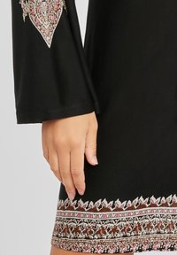 Prenda negra de manga larga, adornada con patrones ornamentales en tonos de rosa y marrón en el dobladillo y las mangas, con una textura de tela suave.