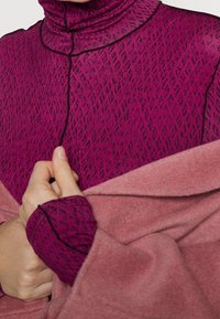 Long-sleeve top in deep magenta with a textured diamond pattern, featuring a high funnel neck and contrasting black trim. Soft pink draped fabric accent.