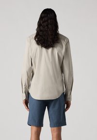 Chemise longue à manches longues beige clair avec des poignets à bouton, ourlet incurvé et texture douce, associée à un short bleu foncé arrivant au genou.