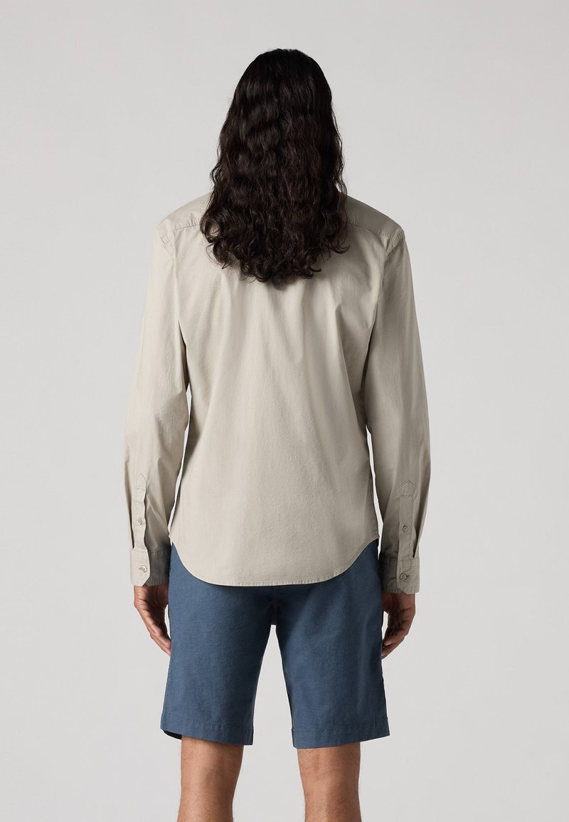 Hellbeige Langarmhemd mit knöpfbaren Manschetten, geschwungenem Saum und weicher Textur, kombiniert mit knielangen dunkelblauen Shorts.