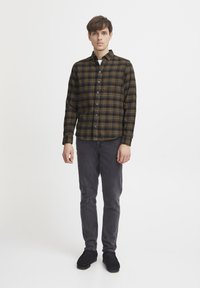 Camisa de botones en un patrón de cuadros verde oscuro y amarillo, de manga larga, con botones negros; combinada con pantalones gris oscuro y zapatos negros.