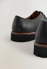 Chaussures en cuir noir avec une finition texturée, forme arrondie et semelle épaisse et dentelée. Présentent un accent marron autour du bord de la semelle.