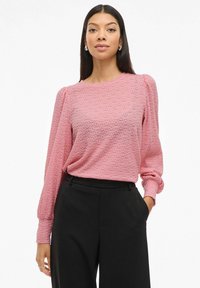 Blusa rosa texturizzata con maniche lunghe e gonfie, scollatura rotonda, indossata con pantaloni neri. Il tessuto presenta un motivo decorativo.