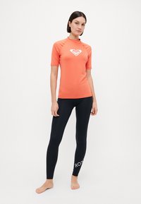 Korall kortermet rash guard med en hvit logo på brystet, sammen med svarte tights med hvit "ROXY"-tekst på underbenet.