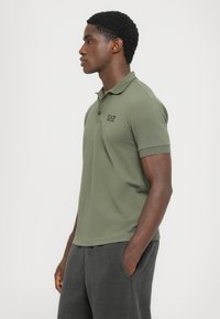 Olive green polo shirt with short sleeves and a classic collar, featuring a small black logo on the chest, made of soft fabric.