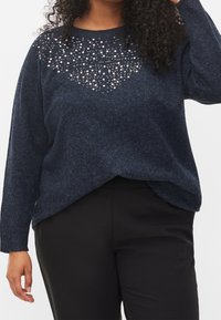 Marinblå stickad tröja med en texturerad design och pärlliknande detaljer på övre framsidan, i avslappnad passform, tillsammans med svarta byxor.